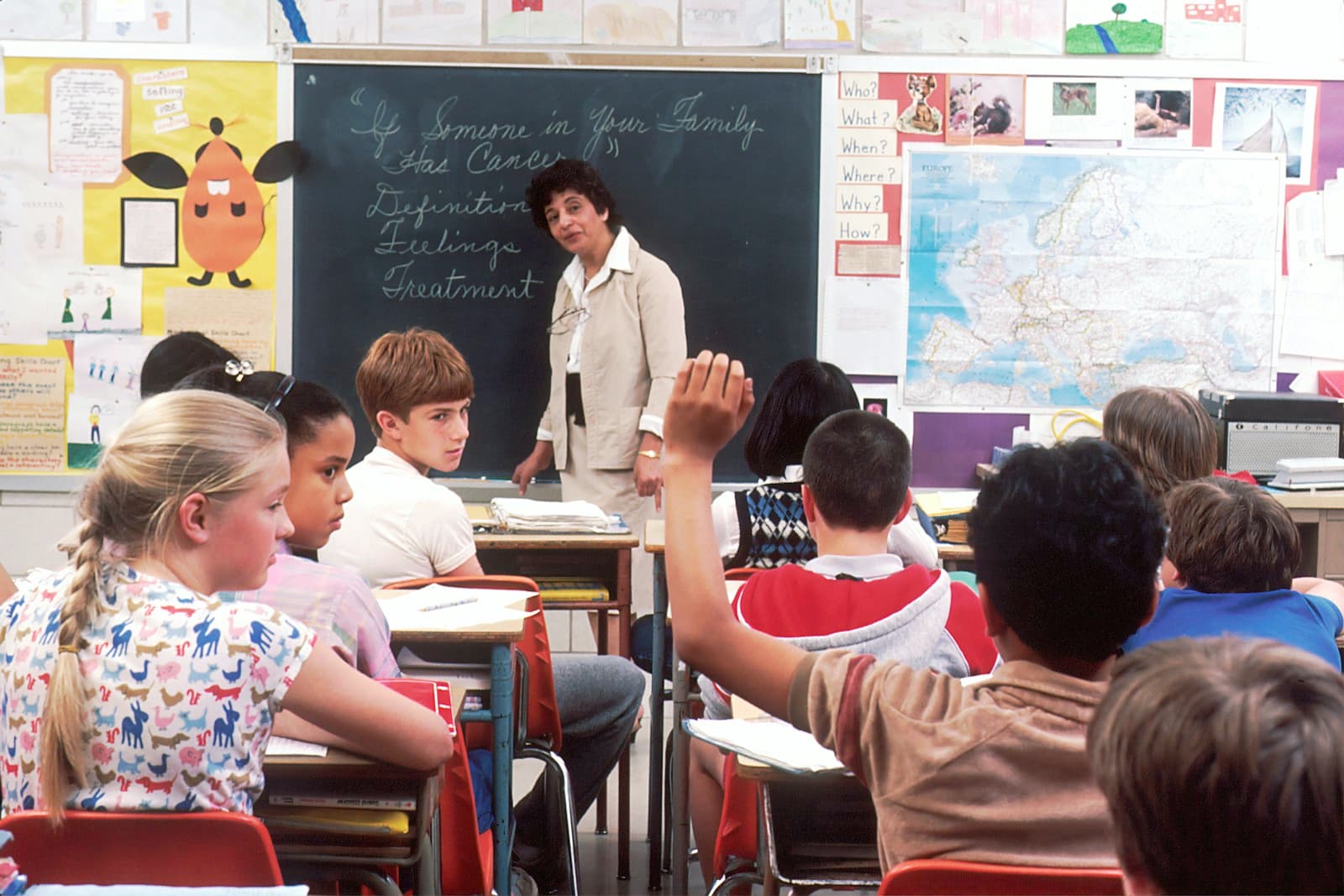 a teacher at a blackboard in front of a classroom of pupils, one with a hand raised