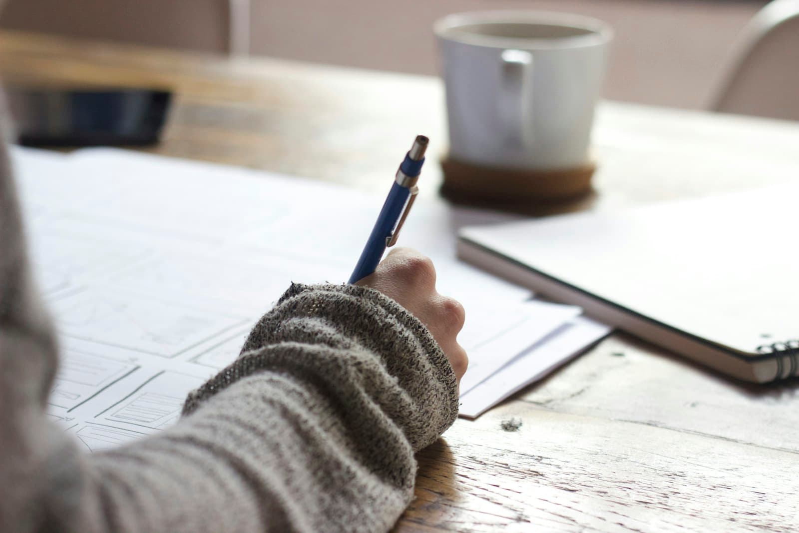 a hand writing in a notebook at a wooden desk beside a mug of coffee