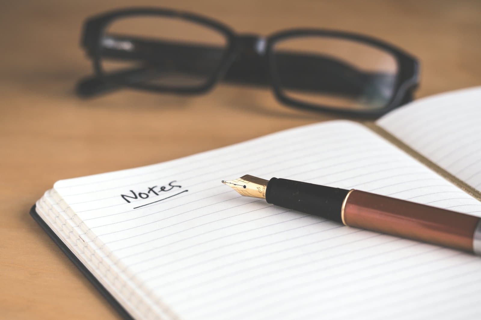 an open notebook with the word Notes handwritten on it, a fountain pen and glasses resting on the page